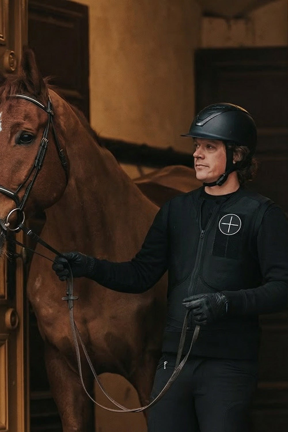 Person in equestrian gear standing next to a brown horse indoors.